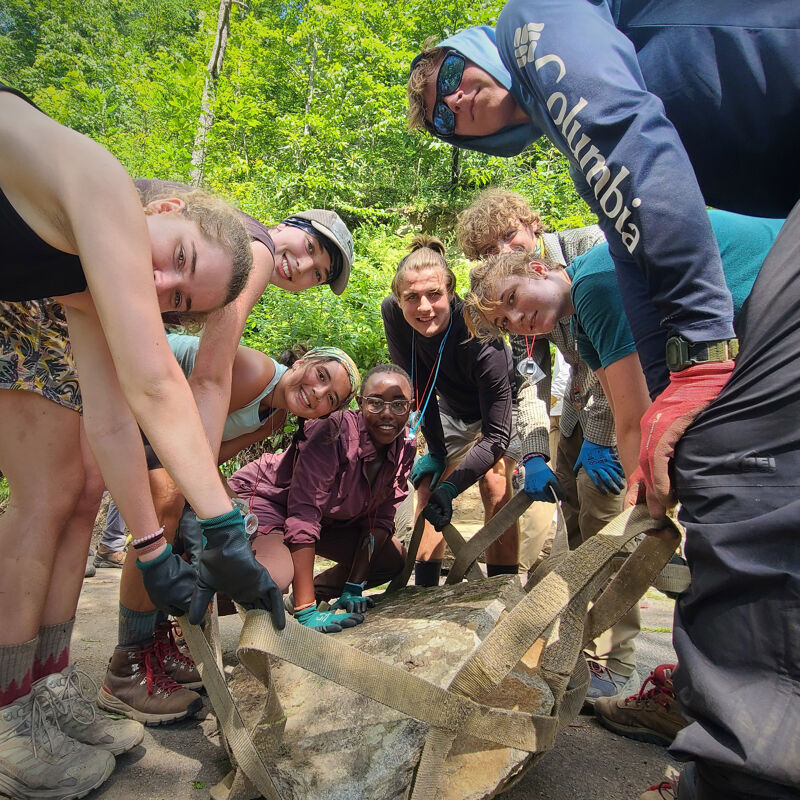 A group of about eight young people are gathered around a large rock, seemingly working together to move it. They are outdoors, surrounded by trees and greenery, suggesting they might be on a trail or in a natural area. Most of them are wearing gloves and casual clothing, and some are using straps to help lift or maneuver the rock. They appear to be collaborating on some kind of outdoor project or activity.